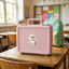 A pink hard-shell lunch box with a Maltese dog sticker sits on a wooden school desk, with a mint green and yellow backpack in the blurred background of a classroom.