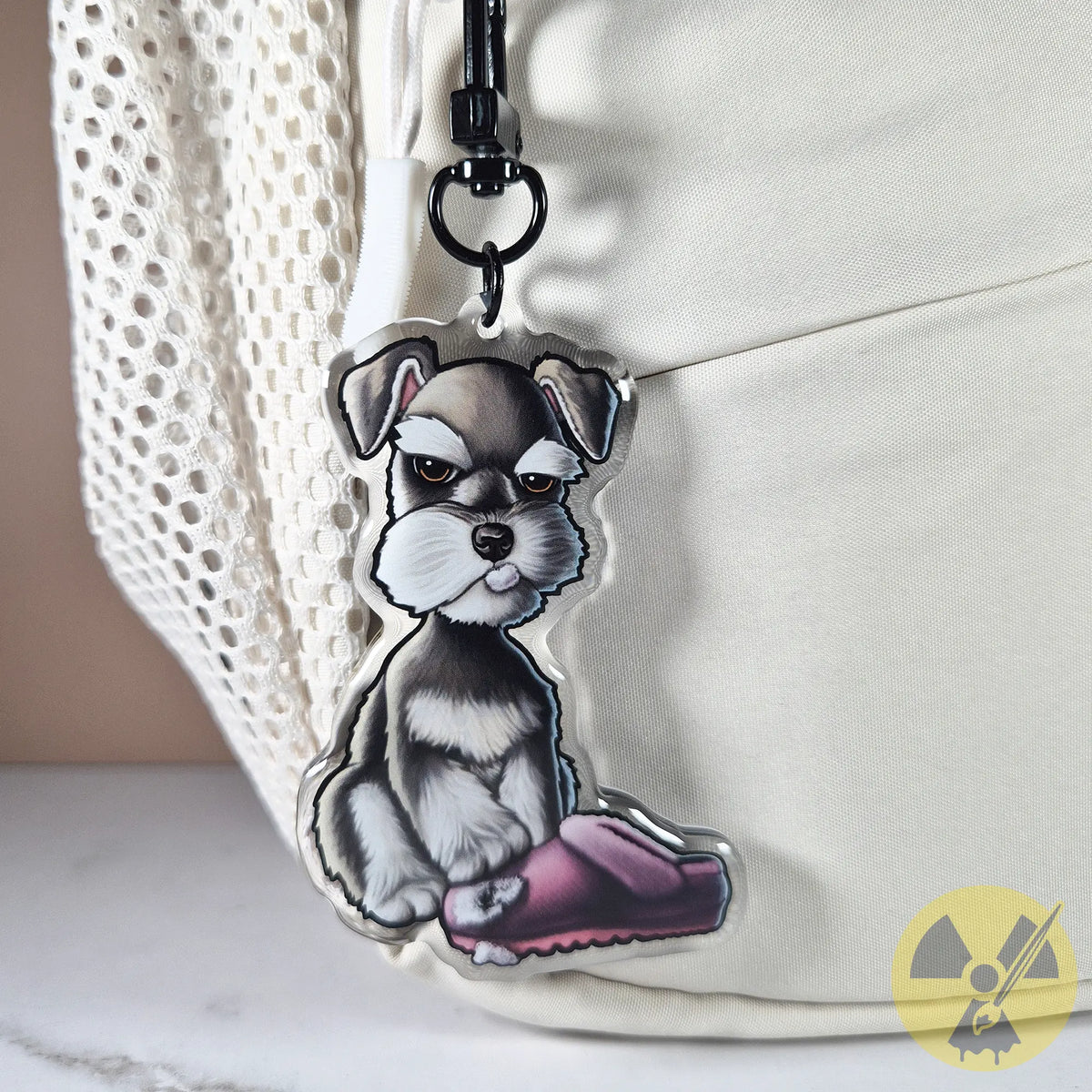 Photo of a keychain of a miniature schnauzer puppy with a chewed pink slipper hanging on a white textured backpack.