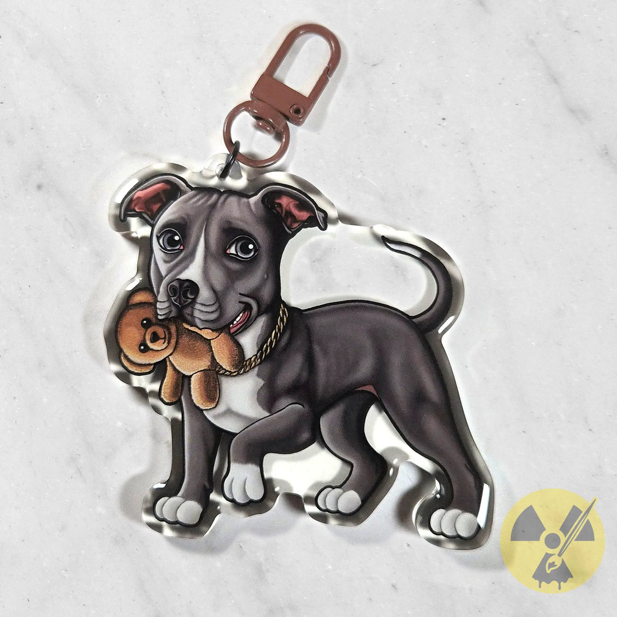 A custom acrylic keychain featuring an illustrated gray and white pitbull dog holding a brown teddy bear, attached to a rose gold metal clasp, shown on a light gray marble background.