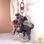 A custom acrylic keychain featuring a cartoon illustration of a gray pitbull dog holding a brown teddy bear, attached to a red metal clasp and shown hanging on a white bag.