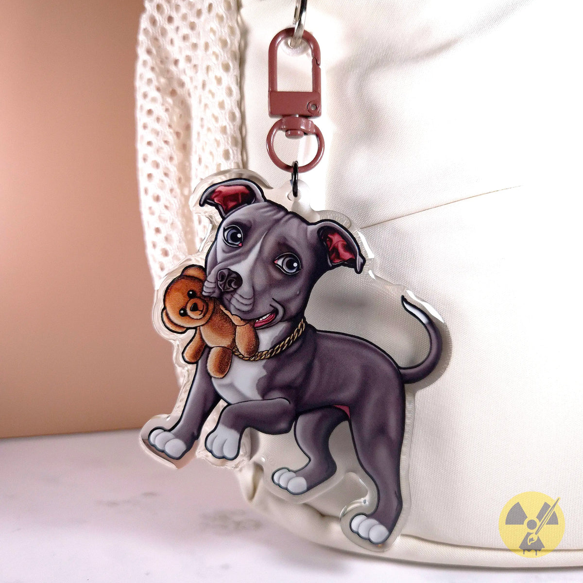 A custom acrylic keychain featuring a cartoon illustration of a gray pitbull dog holding a brown teddy bear, attached to a red metal clasp and shown hanging on a white bag.