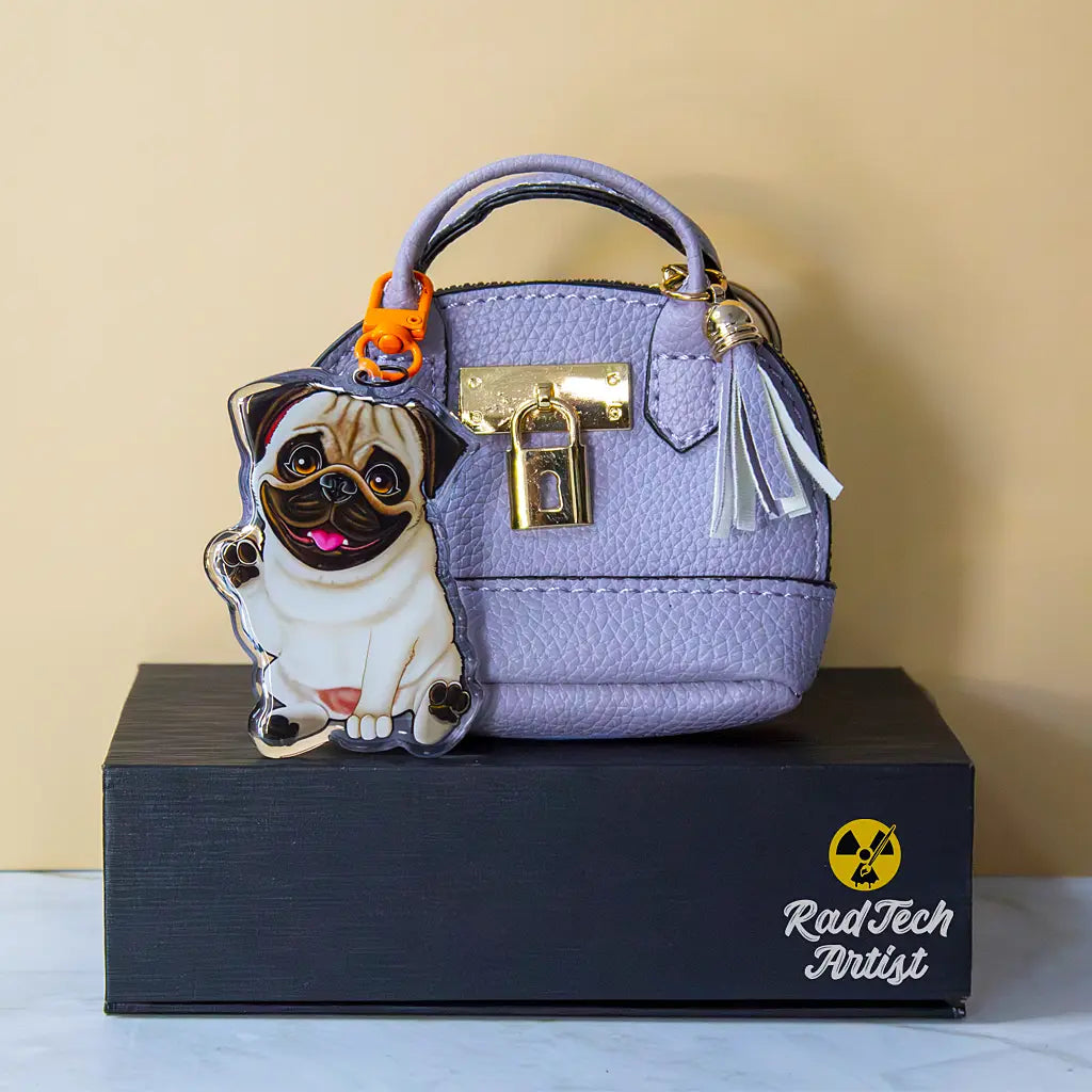 Acrylic pug keychain with epoxy finish, featuring a happy tan pug with dark muzzle and bright eyes. Attached with an orange lobster clasp to a lavender mini purse with gold lock hardware and a tassel, displayed on a black box with the Rad Tech Artist logo.