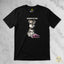 A black short-sleeve graphic t-shirt laid flat on a light gray marble background. The shirt features a cartoon illustration of a salt and pepper Schnauzer puppy sitting by a pink shoe, with the text 'CONSIDER US EVEN.