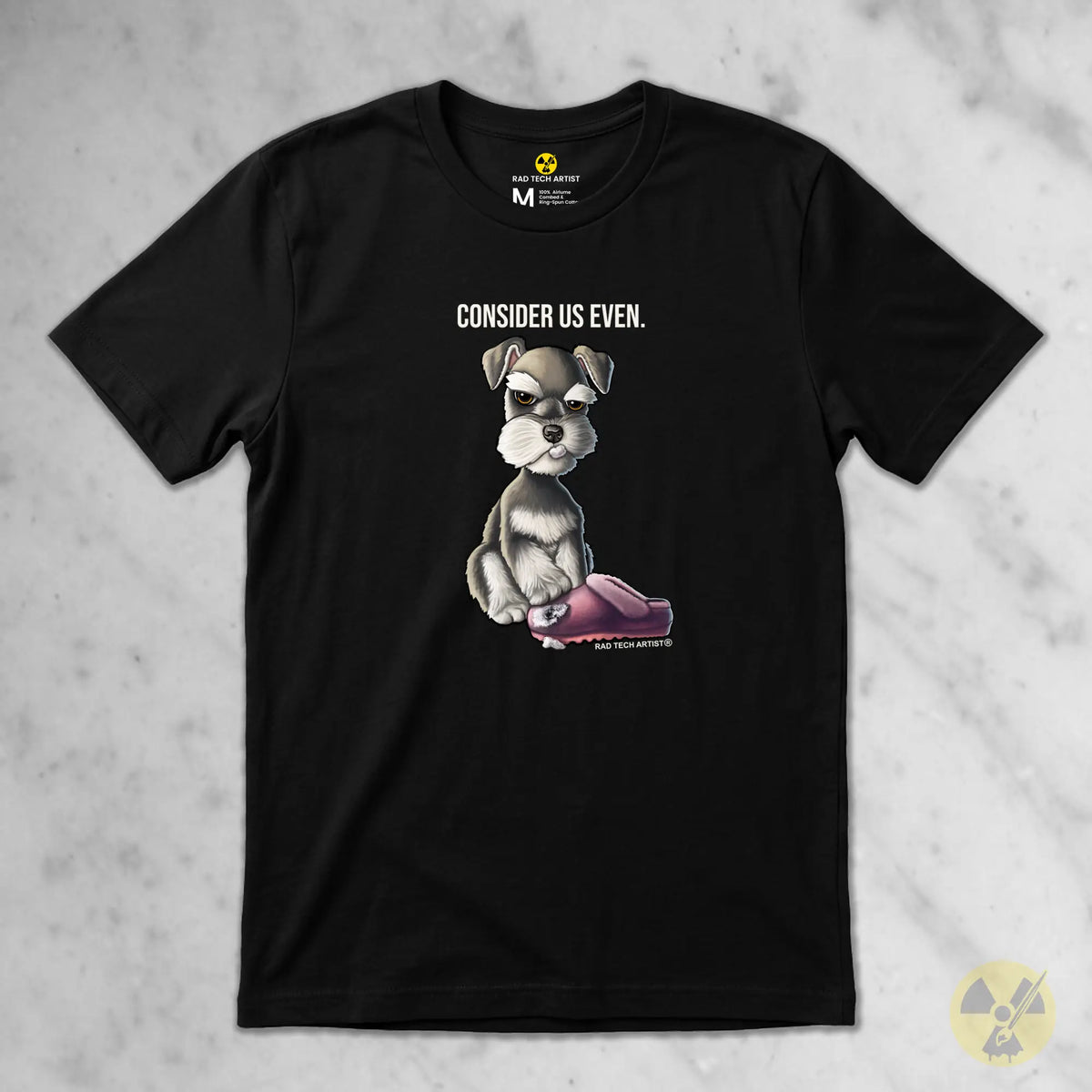 A black short-sleeve graphic t-shirt laid flat on a light gray marble background. The shirt features a cartoon illustration of a salt and pepper Schnauzer puppy sitting by a pink shoe, with the text 'CONSIDER US EVEN.