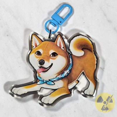 A die-cut acrylic keychain charm featuring a cartoon Shiba Inu dog wearing a blue bandana, attached to a bright blue metal clasp, photographed on a light marble surface.