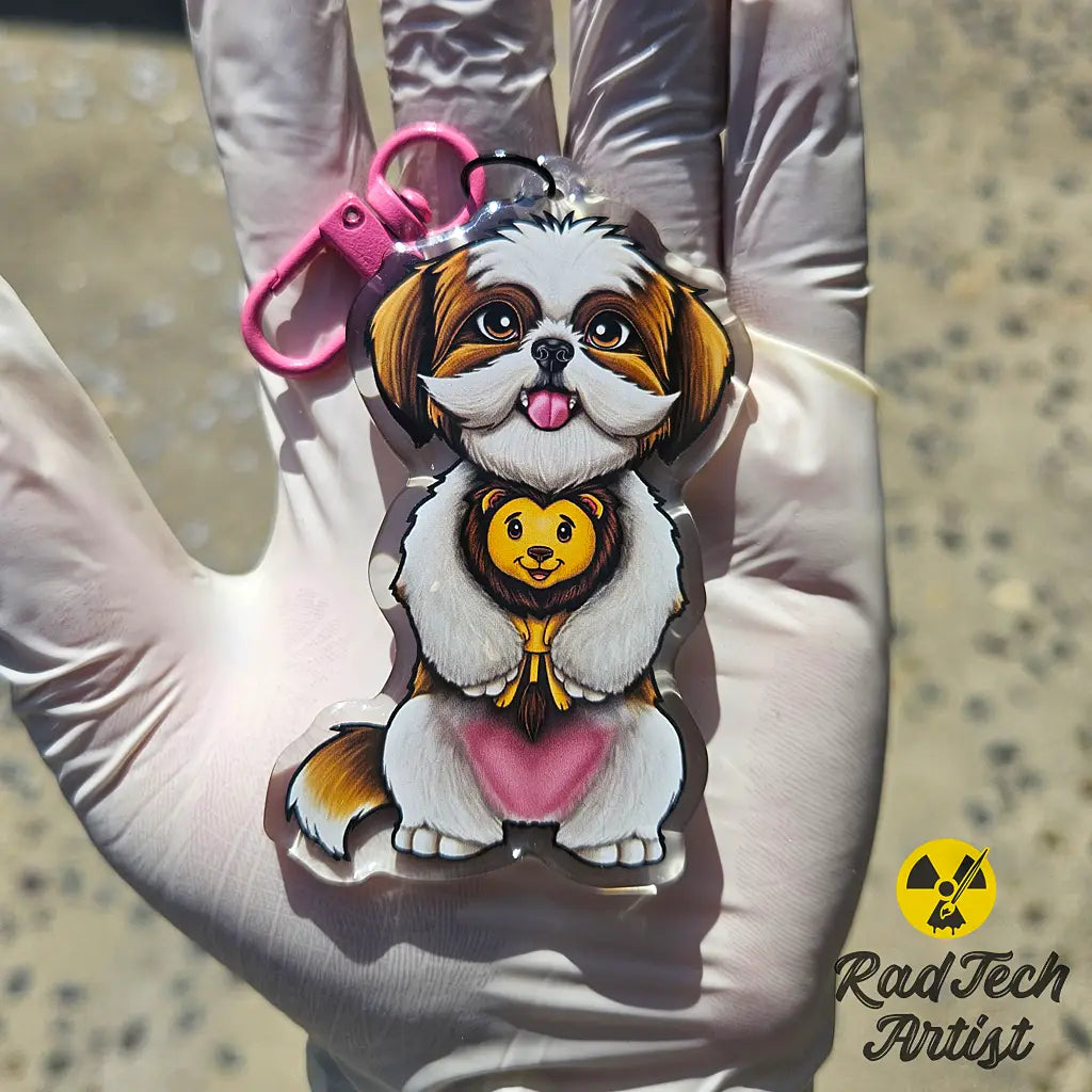 Shih Tzu acrylic keychain with epoxy finish, featuring a fluffy white and gold pup holding a plush lion toy. Attached to a pink lobster clasp and held in a gloved hand under sunlight, with the Rad Tech Artist logo in the corner.
