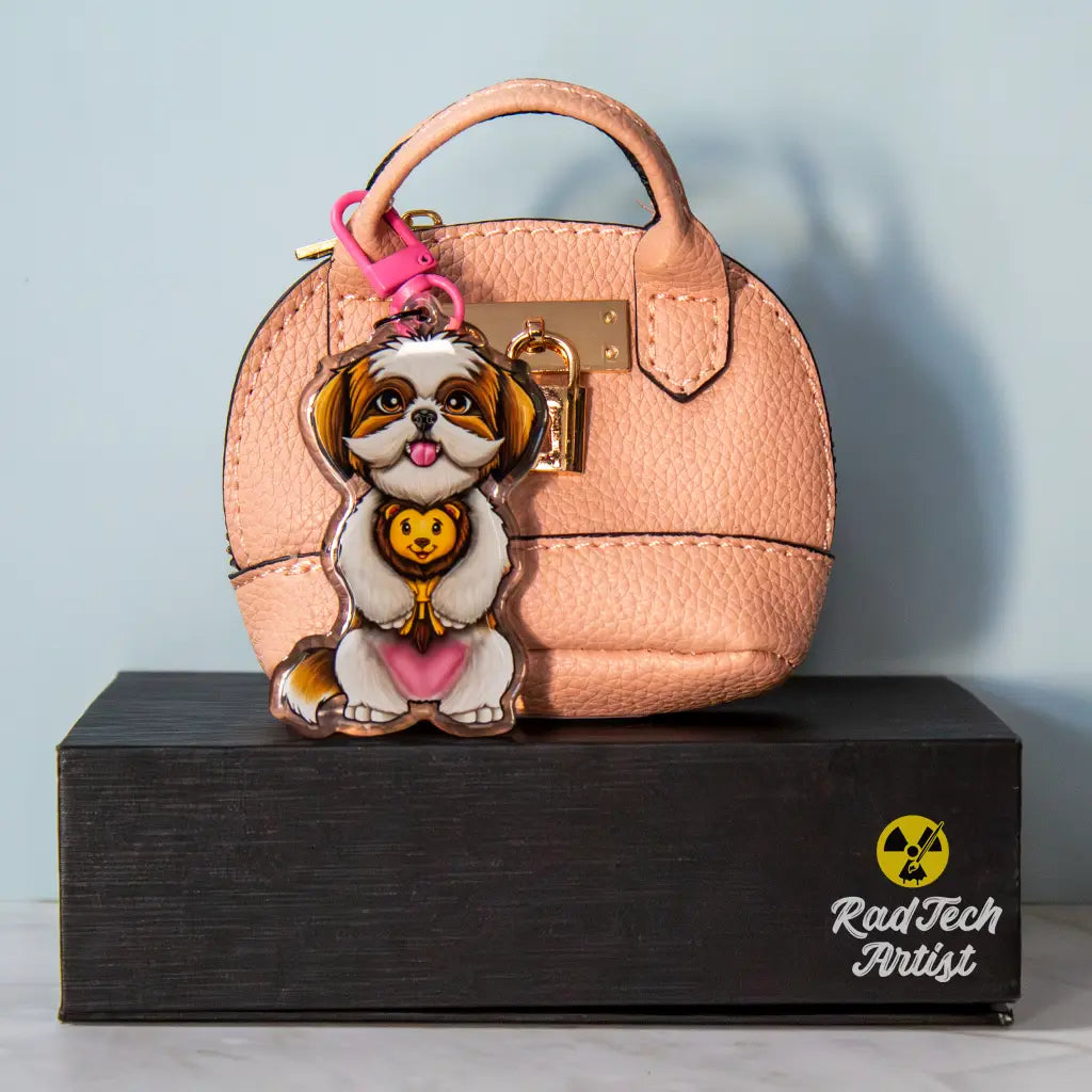 Acrylic Shih Tzu keychain with epoxy finish, featuring a white and gold dog holding a plush lion toy. Attached with a pink lobster clasp to a textured pink mini purse with gold lock hardware, displayed on a black box with the Rad Tech Artist logo.