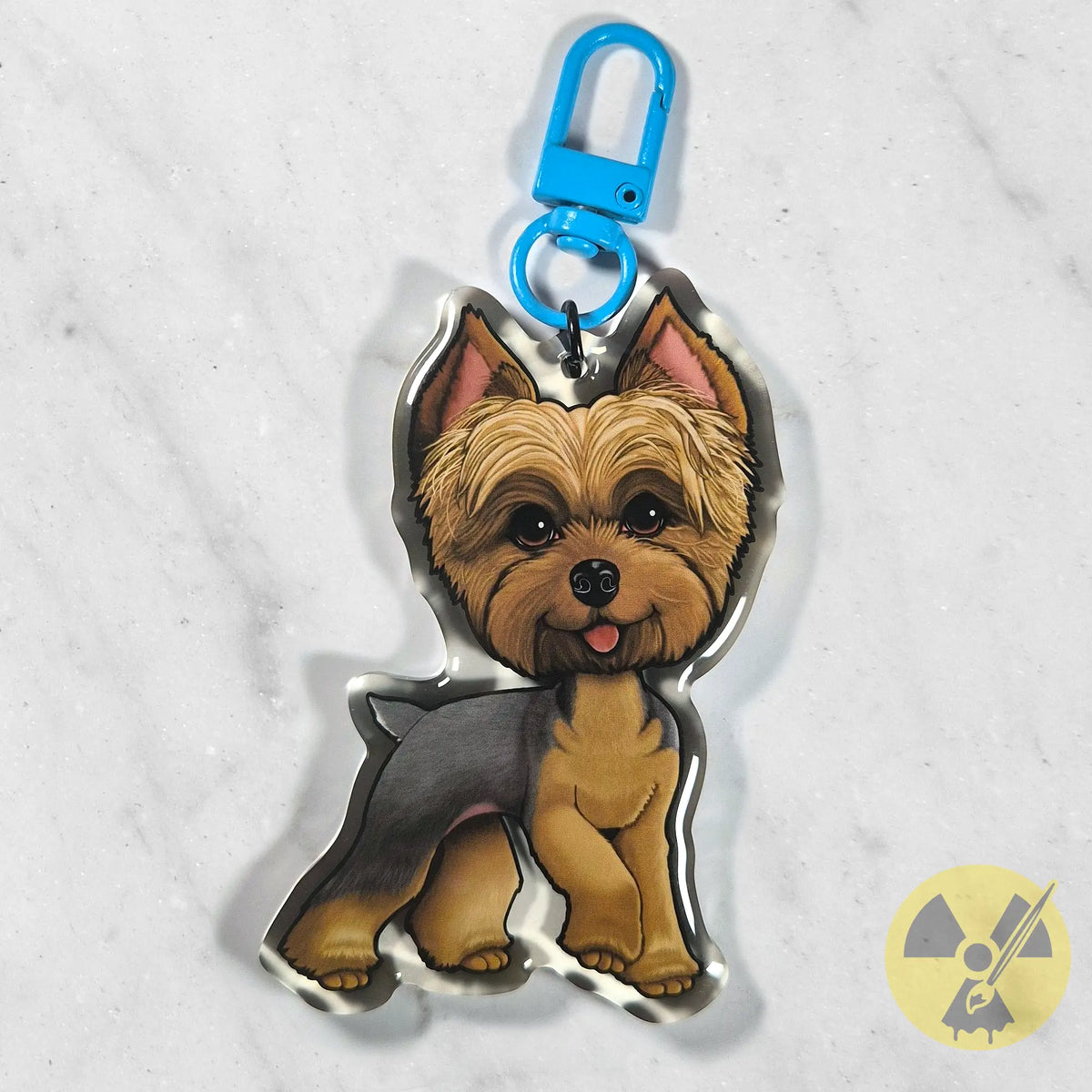 A custom acrylic keychain shaped like a Yorkshire Terrier dog, featuring a bright blue metal clasp at the top. The dog illustration has tan and gray fur with pink inner ears and a small pink tongue.