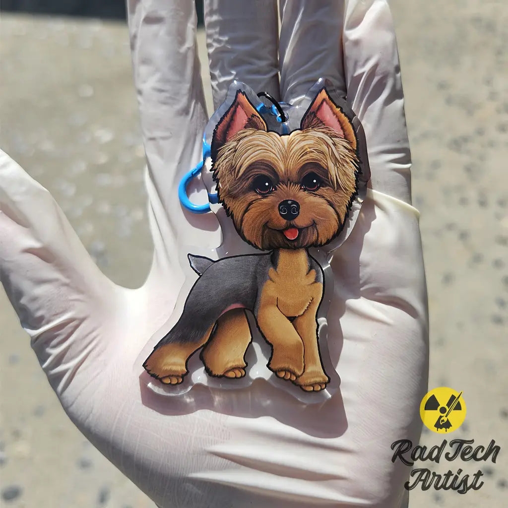 Yorkshire Terrier acrylic keychain with epoxy finish, featuring a blue and gold Yorkie standing proudly. Attached to a blue lobster clasp and held in a gloved hand under sunlight, with the Rad Tech Artist logo in the corner.