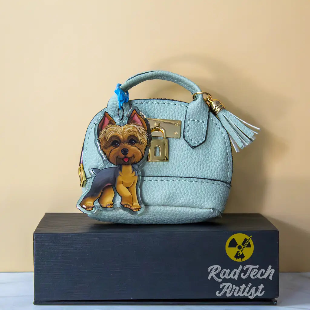 Yorkshire Terrier acrylic keychain with epoxy finish, showing a blue and gold Yorkie standing proudly. Attached to a blue lobster clasp and clipped to a mint mini purse with gold hardware, displayed on a black box with the Rad Tech Artist logo.