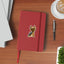 Mock-up image of a blue and gold Yorkshire Terrier sticker on the cover of a red notebook. The Yorkie has a confident expression and silky coat, sitting upright with perked ears. The notebook is placed on a wooden desk alongside a black pen, a latte, and a corner of a potted plant.