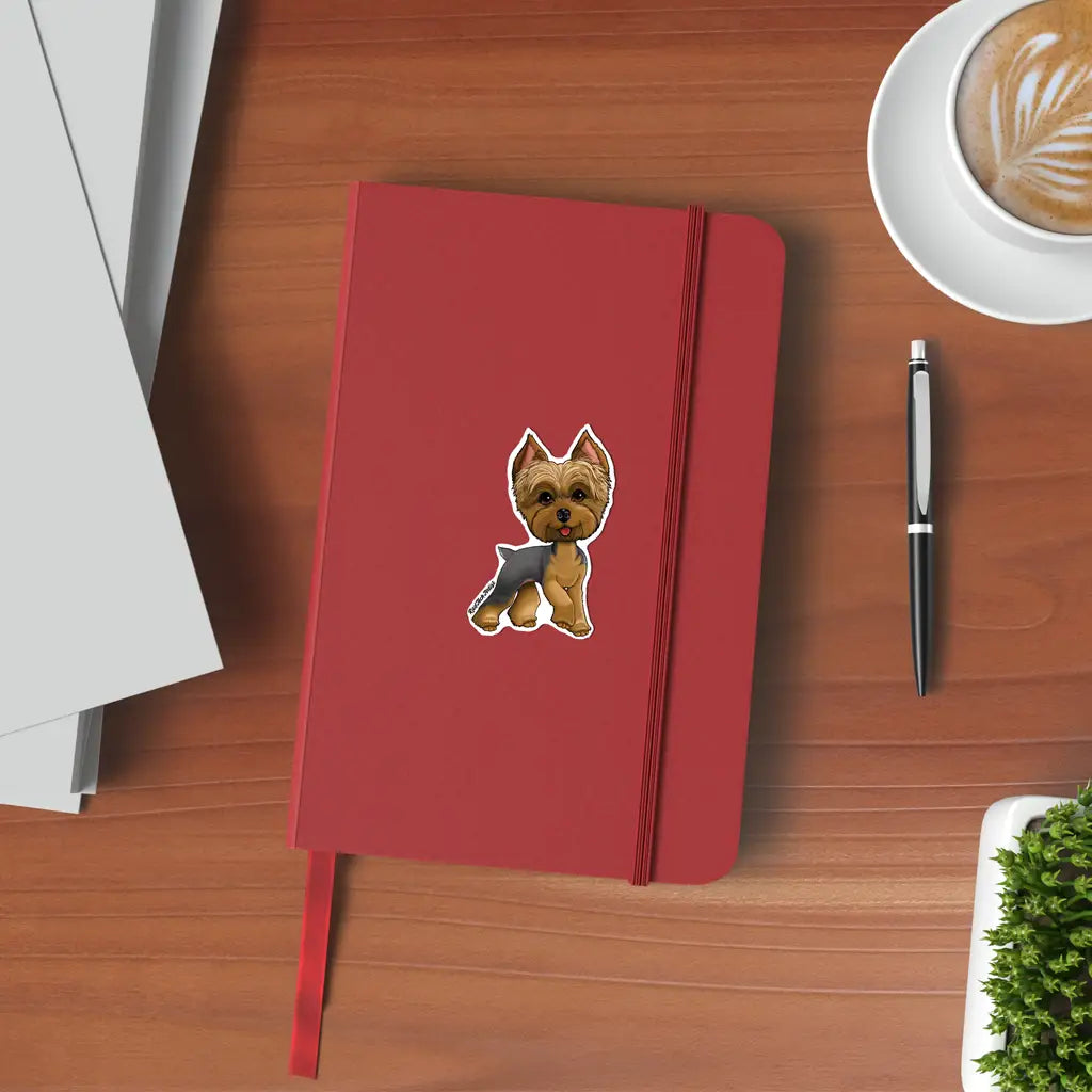 Mock-up image of a blue and gold Yorkshire Terrier sticker on the cover of a red notebook. The Yorkie has a confident expression and silky coat, sitting upright with perked ears. The notebook is placed on a wooden desk alongside a black pen, a latte, and a corner of a potted plant.