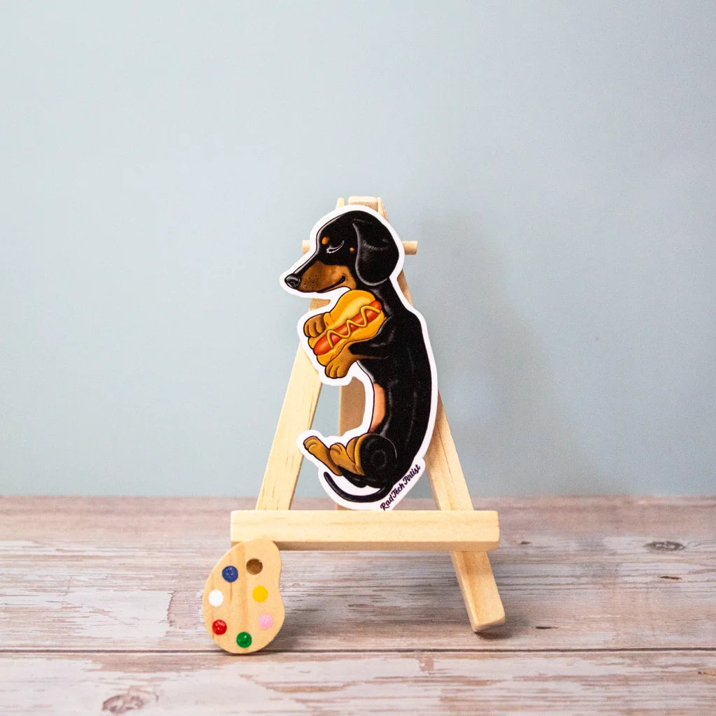 Vinyl sticker of a black and tan Dachshund sitting upright in profile, proudly hugging a plush hot dog toy, displayed on a small wooden easel.