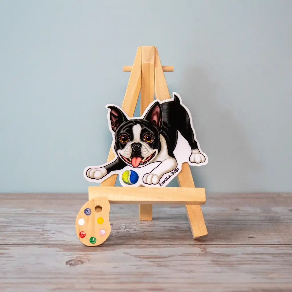 Photo of a black and white Boston Terrier sticker with a playful expression and tennis ball, displayed on a mini wooden easel. A small paint palette prop is placed in front on a rustic wood surface.