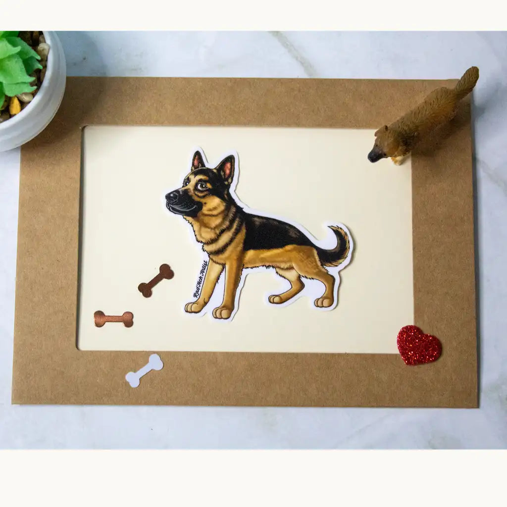 Photo of a black and tan German Shepherd vinyl sticker placed on a bordered card with bone confetti, a red glitter heart, and a miniature dog figure.