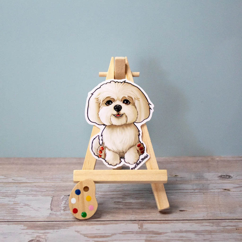 Vinyl sticker of a fluffy white Maltese sitting sweetly on a mini easel, with soft fur and a gentle smile – charming toy breed dog sticker with a calm expression.