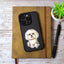 Photo of a Maltese dog sticker with fluffy white fur on a black iPhone case, resting on a brown notebook beside a pen and a small potted plant.