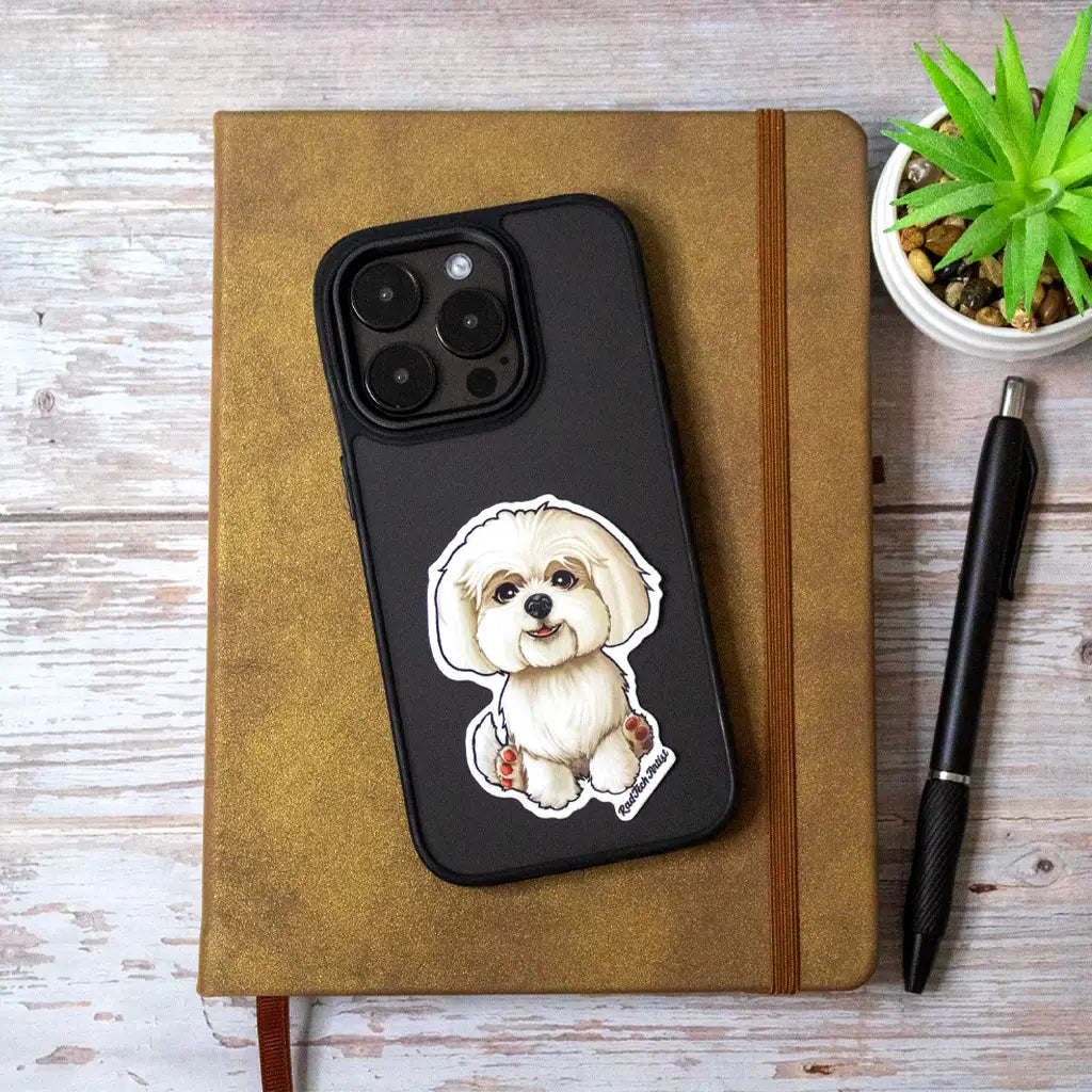 Photo of a Maltese dog sticker with fluffy white fur on a black iPhone case, resting on a brown notebook beside a pen and a small potted plant.