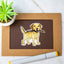 Actual photo of a Yellow Labrador Retriever vinyl sticker displayed on a brown and black bordered card. The Labrador has a friendly expression and is holding a large stick in its mouth. A potted succulent plant and a dual-tip marker sit nearby for decoration.