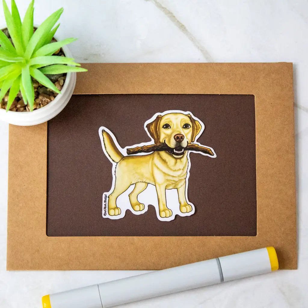 Actual photo of a Yellow Labrador Retriever vinyl sticker displayed on a brown and black bordered card. The Labrador has a friendly expression and is holding a large stick in its mouth. A potted succulent plant and a dual-tip marker sit nearby for decoration.
