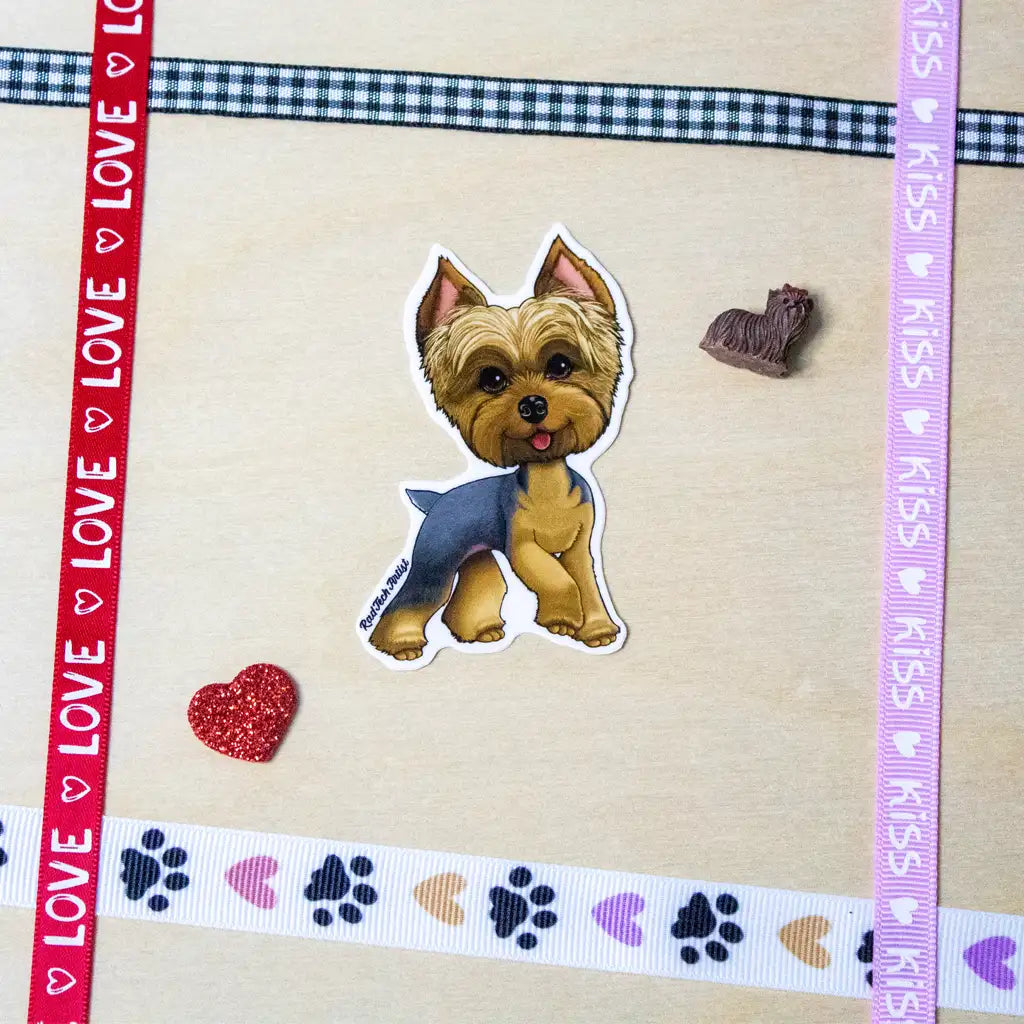 Photo of a blue and gold Yorkshire Terrier vinyl sticker with a sassy expression, placed on a beige surface decorated with colorful ribbon borders, paw prints, hearts, and a small Yorkie figurine.