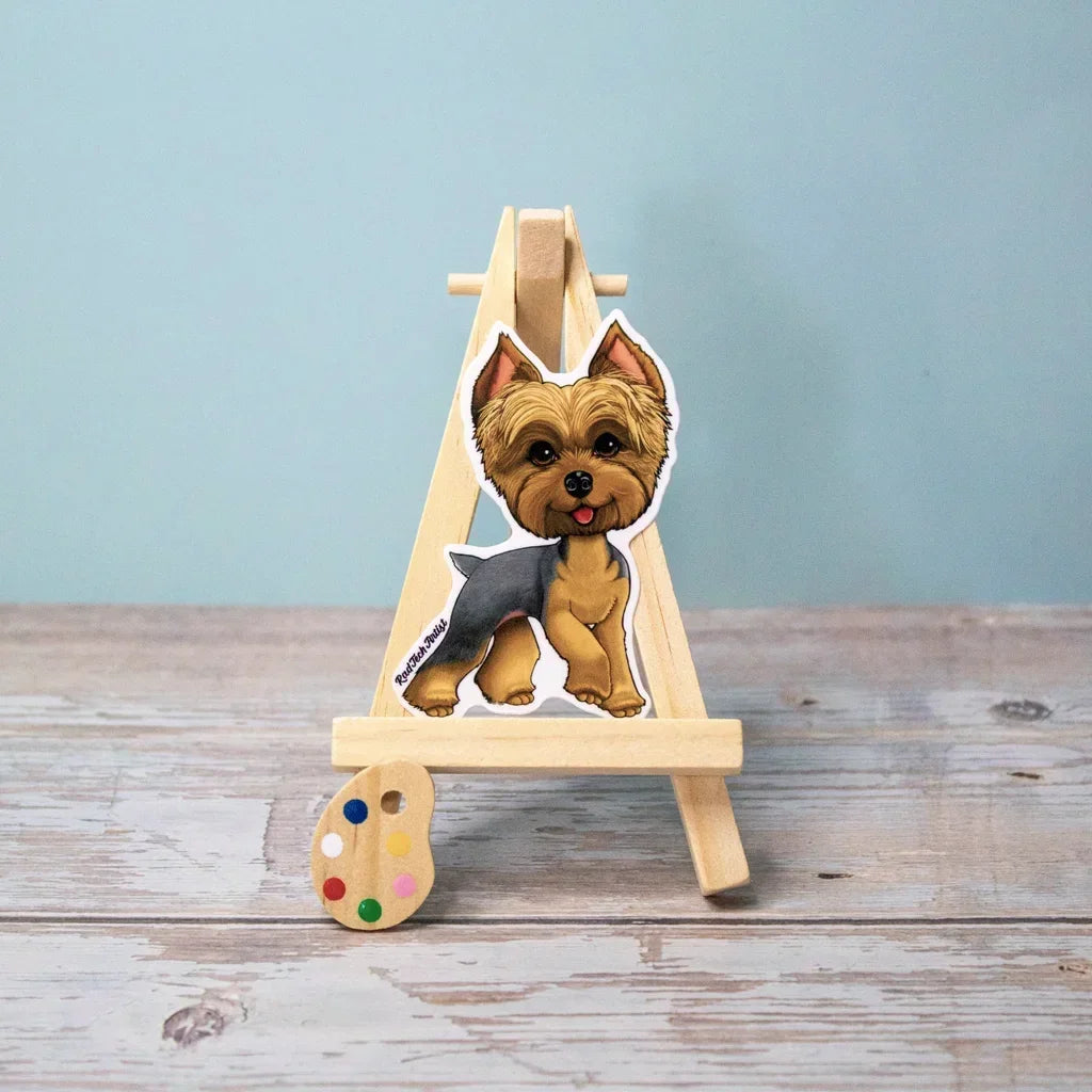 Photo of a blue and gold Yorkshire Terrier vinyl sticker displayed on a small wooden easel with a tiny paint palette, set against a light blue wall and rustic wooden surface.
